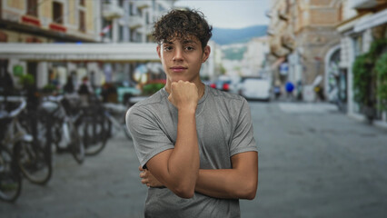 Fototapeta premium Teenage boy with fist under chin and arm crossed, posing on a street with parked bicycles and buildings in view; thoughtful introspection youth.
