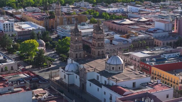 Aerial drone footage captures Durango Cathedral with twin bell towers and ornate blue dome in historic downtown Mexico. Colorful colonial buildings and tree-lined plaza surround the baroque church.