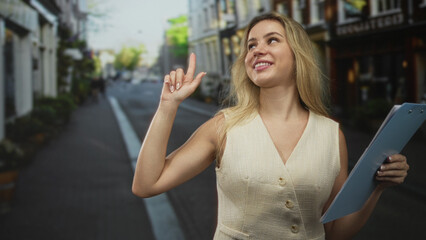 Fototapeta premium Young woman points finger upward on street while holding clipboard and smiling; confidence leadership assertiveness.