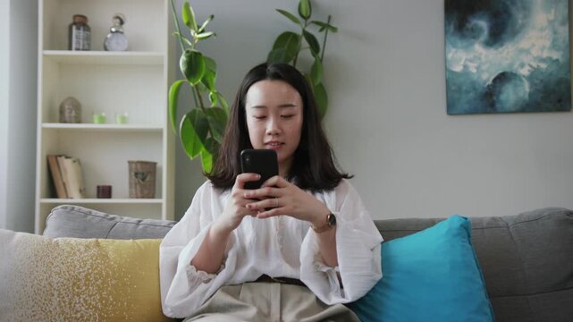 Shooting of nice smiling young woman looking at screen of smartphone, messaging. Lovely pet jumping on couch, wagging tail.