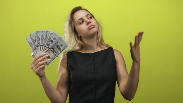 Young blonde woman holding a fan of dollar bills with hand raised fanning cash in a green studio; indifference wealth.