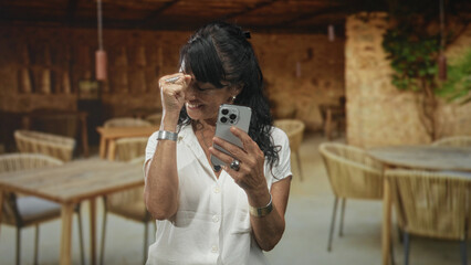 Obraz premium Woman senior hispanic holding smartphone and pumping fist while smiling in building dining space into camera; joy celebration.