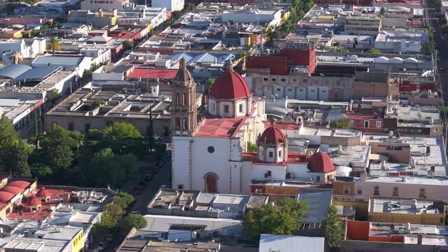 Aerial drone footage captures stunning colonial cathedral with distinctive red domes and baroque bell tower rising above the urban landscape of Durango Mexico. Green trees surround the historic.