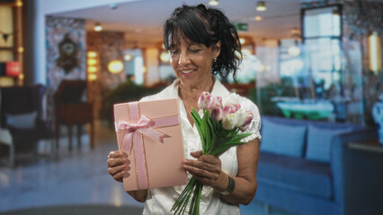 Obraz premium Woman holding pink gift box and tulips, smiling and presenting bouquet in a hotel lobby building; celebration joy.