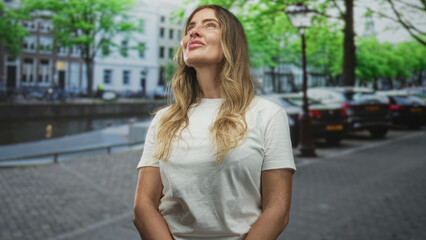 Fototapeta premium Middleaged hispanic woman face smiling and looking up on street by a canal with parked cars, lamp post, trees and riverside buildings; contentment.