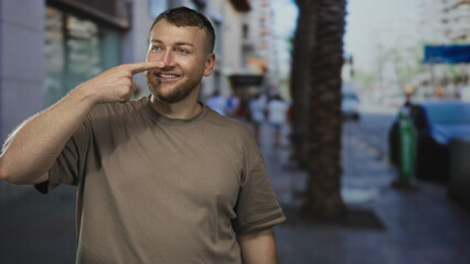 Fototapeta premium Man points finger to nose on a city street, bearded and wearing a brown tshirt, smiling; playful curiosity.