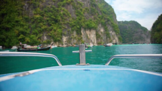 POV from blue speedboat bow with chrome cleat crossing emerald tropical waters as traditional longtail boats float near dramatic limestone cliffs covered in lush green jungle vegetation in Thailand.