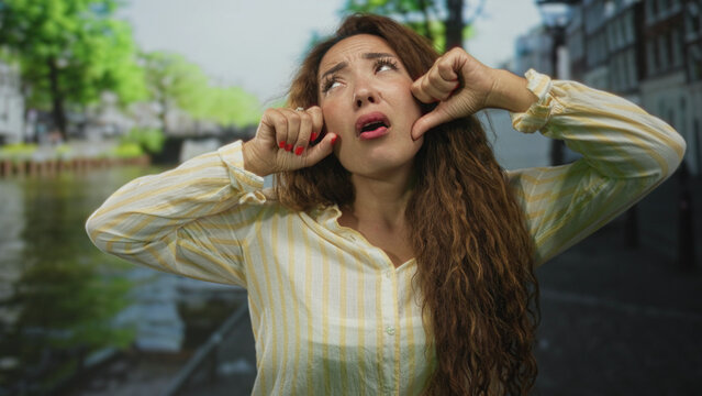 Young woman pulls her cheeks and pouts on a street in amsterdam beside a canal wearing a striped shirt; sulking playful humor.