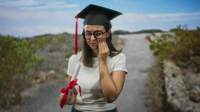 Young hispanic woman wearing graduation cap holding rolled diploma tied with red ribbon touches cheek with hand for toothache on rural street; reflection perseverance pain.