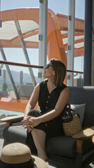 Obraz premium Woman holding sunglasses on cruise terrace lounge chair with straw bag and hat beside her while she sits and reaches toward hat; travel leisure calm.