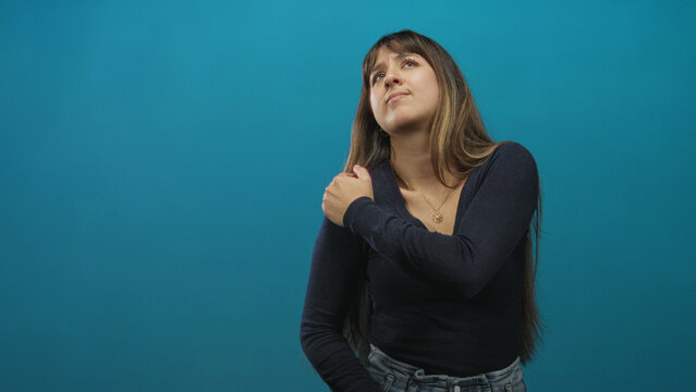 Young woman clutching shoulder, wincing while wearing sweater and jeans on studio backdrop; pain vulnerability recovery.