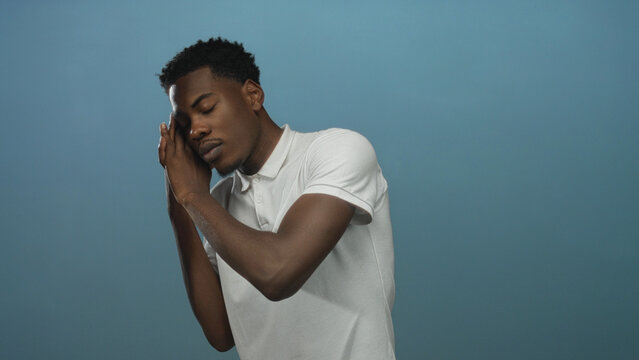 Young african american man sleeping with hands at cheek against pale blue background in studio environment; serenity.