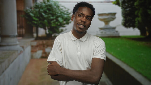 Man wearing sunlit crisp white polo shirt crosses his arms in street by a building under soft morning light; confidence.