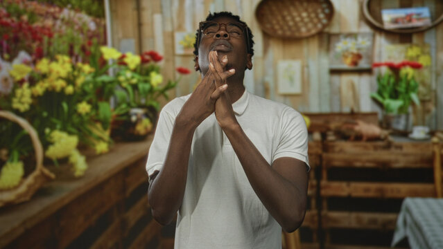 Young man with glasses clasps interlaced hands and looks upward in a bright flower shop stall; hope faith.