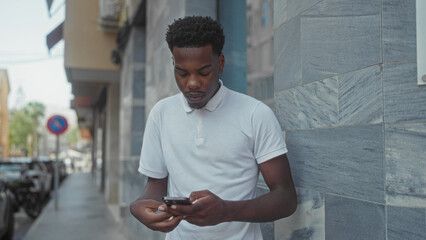 Man in white polo shirt holding smartphone scrolls screen while leaning against stone building on...