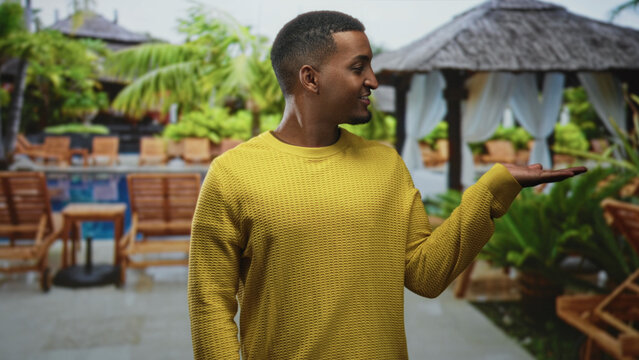 Man presenting palm up at building pool cabana wearing yellow sweater, palm extended in gesture of offering; welcoming calm.