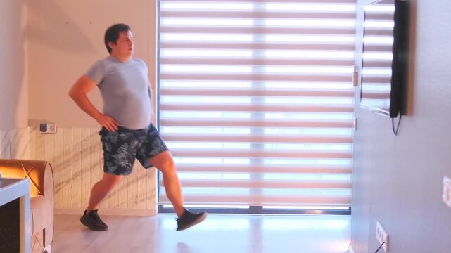 Caucasian man lunges in living room facing TV and horizontal blinds, wearing grey shirt and patterned shorts, standing on hardwood floor, focused home workout session with steady balance