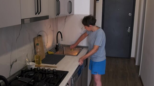 Woman washing metal kitchen sink with sponge and detergent, removing dirt, grease and stains from surface, maintaining hygiene and cleanliness in modern kitchen, daily household routine