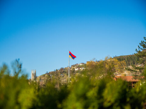 Armenian flag on tall flagpole in the sky over Dilijan, Tavush Province, Armenia. Mountains, and a monument to 50 years of Armenia within the Soviet Union in the background and a bush in foreground