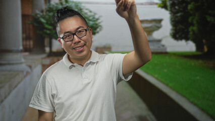 Obraz premium Man points finger upward with hand in building courtyard wearing white polo and glasses, topknot hairstyle visible; playful greeting.