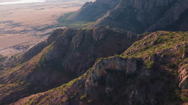 Aerial drone footage captures the rugged rocky peaks and green vegetation of mountainous terrain during golden hour, with dramatic cliffs overlooking vast arid farmland plains below in warm light.