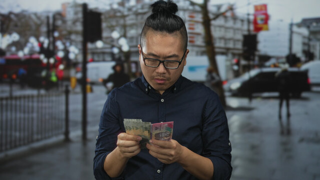 Indonesian man counting rupiah banknotes with both hands on a wet city street near traffic and sidewalk railings, wearing glasses; financial concern.