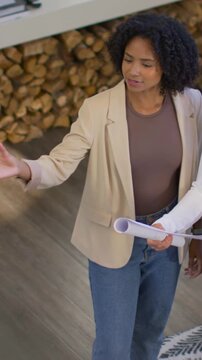Vertical video: Reviewing rolled plans, woman in beige blazer and jeans gesturing left at home