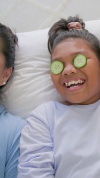 Vertical video: Shot opening on mom laughing with cucumber slices on bed, kid removing them playing