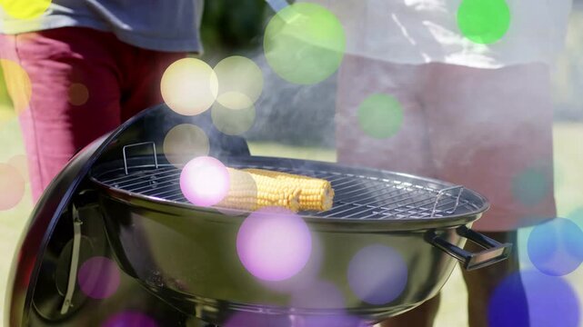 Two men lifting corn with tongs and placing skewer on grill, preparing meal under colorful bokeh