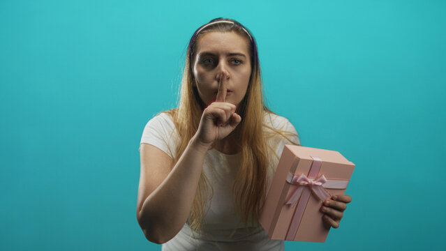 Woman holding a pink present and pressing finger to lips in studio, leaning forward with worried glance and tense expression; secret silence.