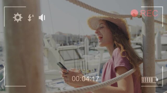 Teen tapping phone at marina reading msg smiling laughing glancing at cam REC overlay framing tech
