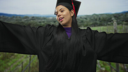 Naklejka premium Young latina woman in black graduation gown and mortarboard with red tassel spreads arms in green countryside field; academic achievement pride.