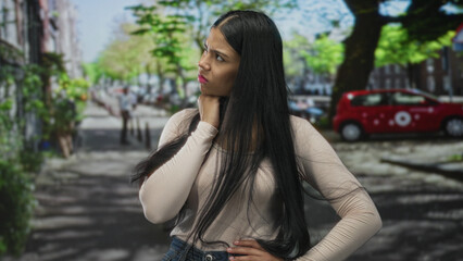 Fototapeta premium Woman standing with hand on chin in pensive gesture on street amid parked cars; reflection introspection contemplation concern.