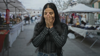 Obraz premium Young latin woman covers mouth with hands on a street market with colorful souvenir stalls visible; surprise.