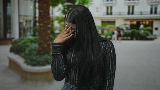 Woman with long black hair and glasses touches her forehead gesture on urban street outside a building; stress.