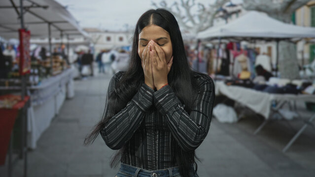 Young latin woman covers mouth with hands on a street market with colorful souvenir stalls visible; surprise.