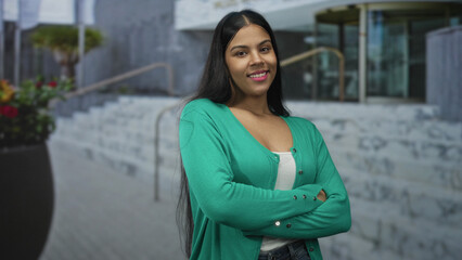 Fototapeta premium Woman stands with arms crossed in green cardigan at modern building entrance; confidence empowerment.