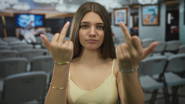 Teen girl standing flips both middle fingers toward camera in spacious gallery building; defiance anger.