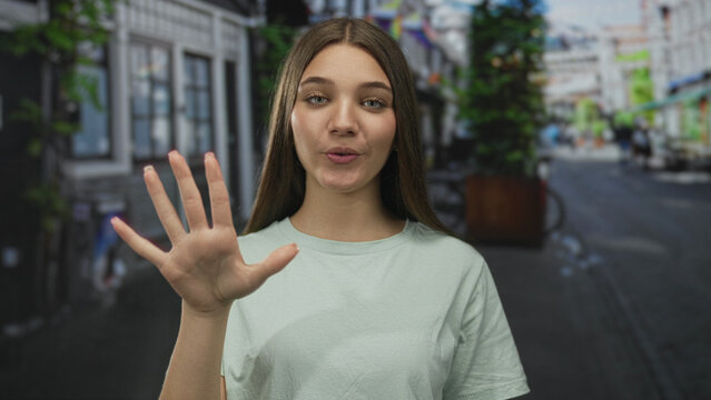 Teenager girl waves hand on bustling city street with storefronts and bicycles in sunny weather; friendly welcome.