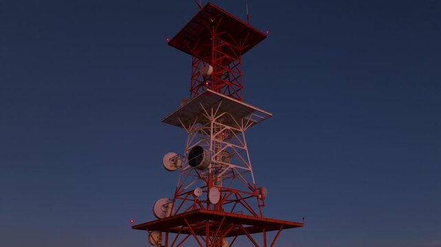 Aerial drone footage orbiting red and white telecommunications tower with satellite dishes and warning lights against a stunning gradient twilight sky over illuminated urban sprawl of Durango, Mexico.