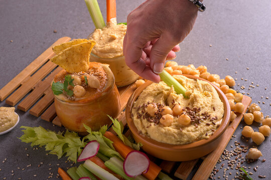 Hand of a woman dipping a fresh vegetable stick into creamy hummus bowl with seeds and chickpeas