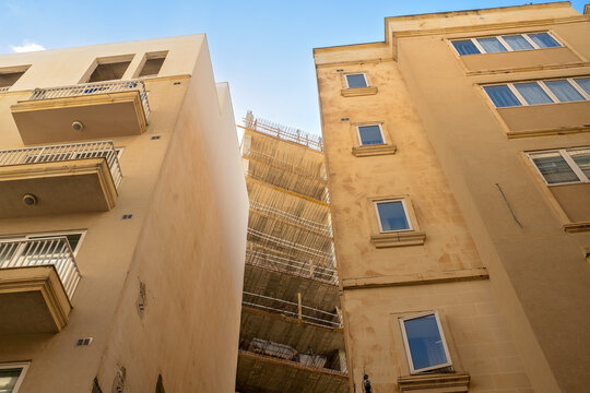 A low-angle view of a new multi-story residential building under construction with concrete floors and steel reinforcement, tightly squeezed between two existing apartment blocks. 