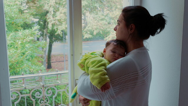 Mother standing by window holding sleeping baby in arms, calm domestic moment expressing tenderness, care, and quiet love in home environment