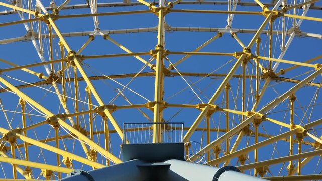 Close up of a giant yellow Ferris wheel spinning against a perfectly clear blue sky. The intricate steel structure slowly rotates