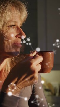 Vertical video: Mature woman lifting cup in kitchen savoring wellness, particles passing over face