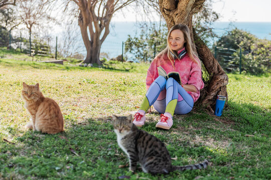Woman reading by a tree in a park with cats