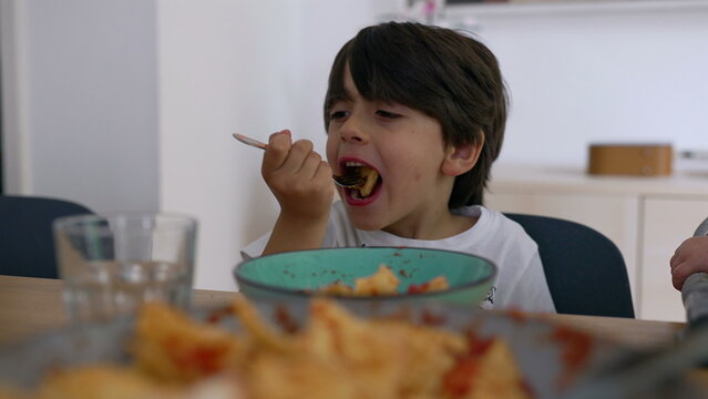 Young boy eating pasta at table, candid moment of appetite and joy during family mealtime in bright home setting
