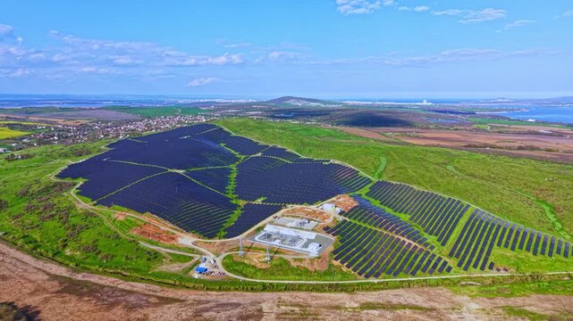 A wide field shows many solar panels installed on a hillside. The area is surrounded by greenery and distant hills under a blue sky. The scene occurs during the day.