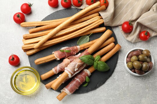 Grissini breadsticks with prosciutto, tomatoes, oil, olives and basil on light table, flat lay