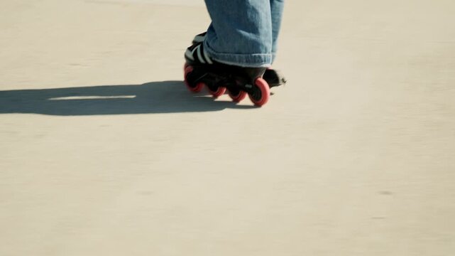 Gimbal tracking close-up of red-wheeled inline skates gliding on sunny concrete promenade. Wide jeans hem, sharp wheel detail, shadow and motion blur below. Dynamic urban active sport and freedom.
